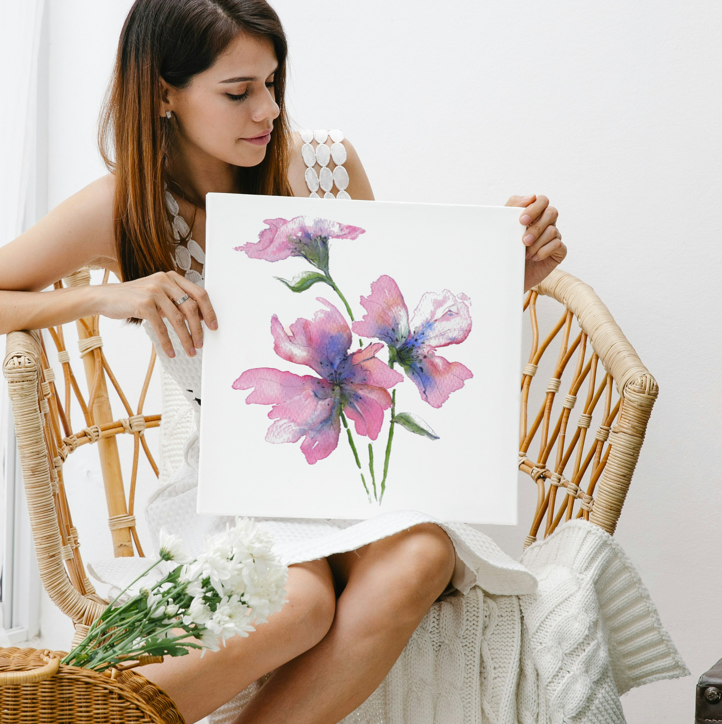 Woman holding a painting of flowers in a wicker chair with plants around.