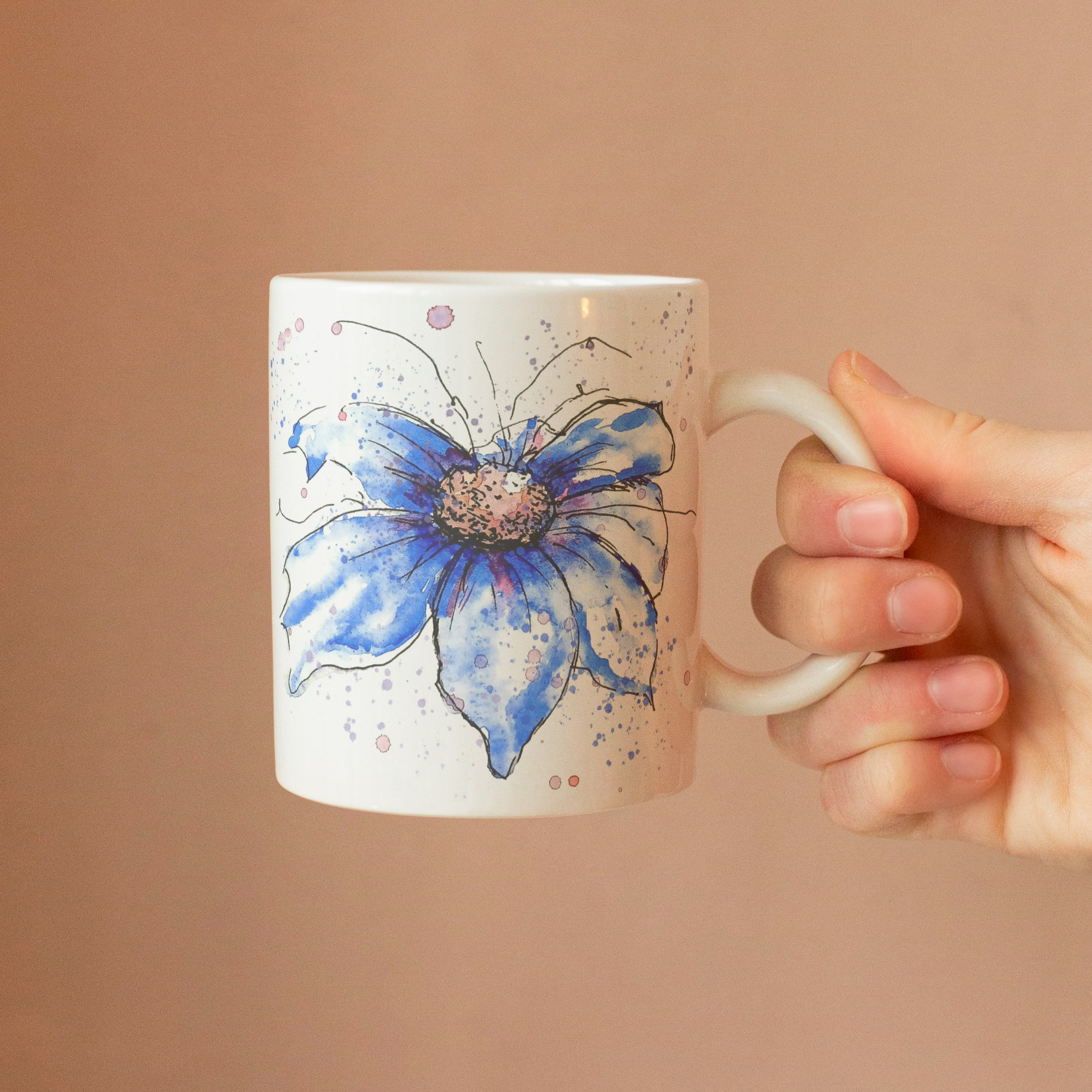 Blue cosmos watercolour mug held in hand against warm peach wall showing comfortable grip