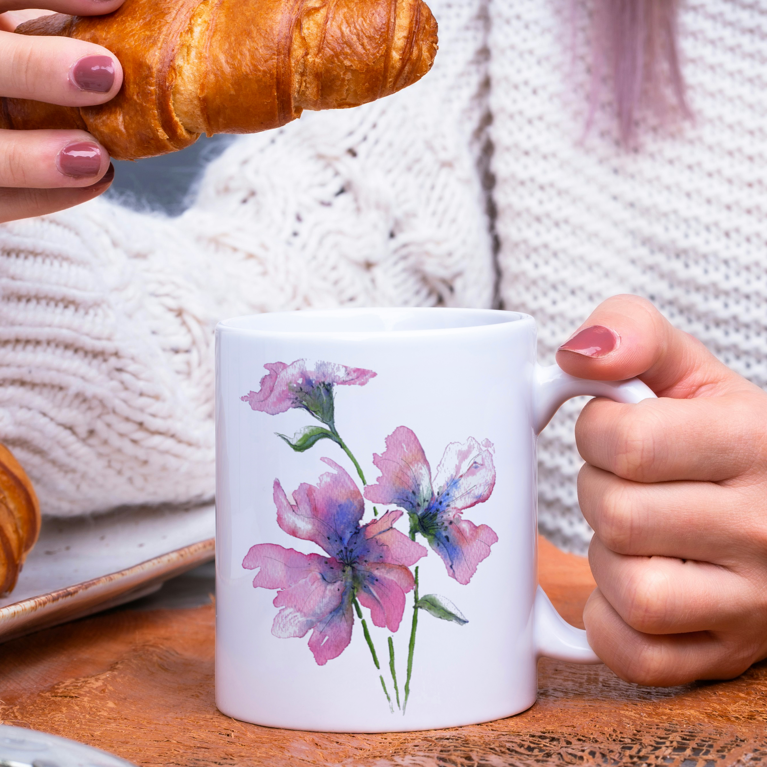 Person holding a mug with floral design and a croissant on a wooden table.